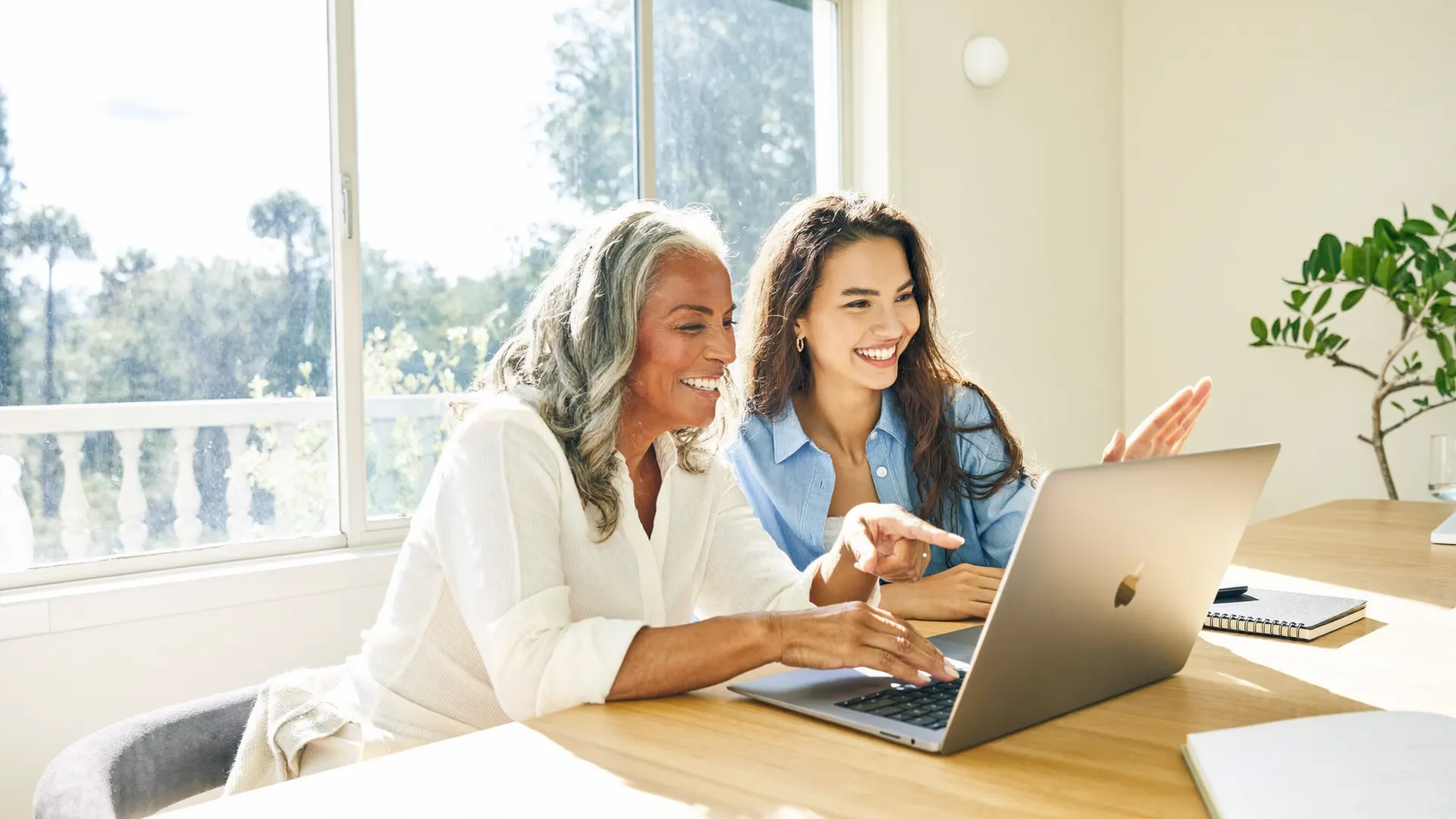 Uma mulher de cabelos grisalhos e uma mulher mais jovem sorriem lado a lado diante de um laptop, apontando juntas para a tela.